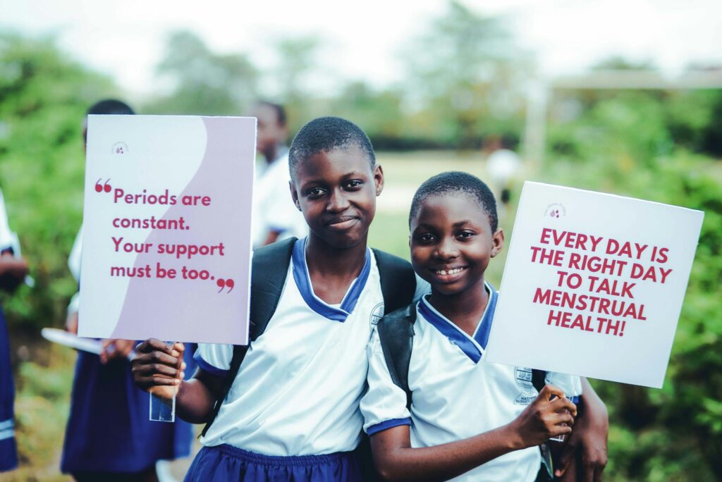 Two students hold signs promoting menstrual health awareness, smiling outdoors.